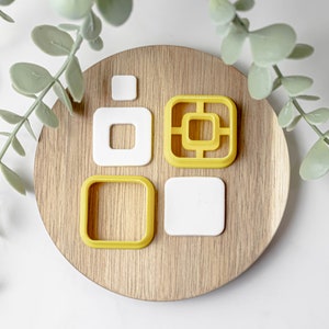 May include: Four cookie cutters on a wooden surface. The cutters are in the shape of squares, with one having a smaller square cut out of the centre. Two cutters are yellow and two are white.