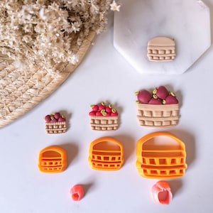 May include: A collection of basket-shaped cookie cutters and basket-themed clay art. The cutters are orange, and the clay art features baskets filled with red berries. A woven basket and dried flowers are in the background.