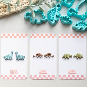 May include: Three cards with pairs of dinosaur-shaped polymer clay earrings. The earrings are on white cards with a pink and white checkered border. The text "Echo Stone" is printed on each card. The earrings are green, brown, and light green with white polka dots.