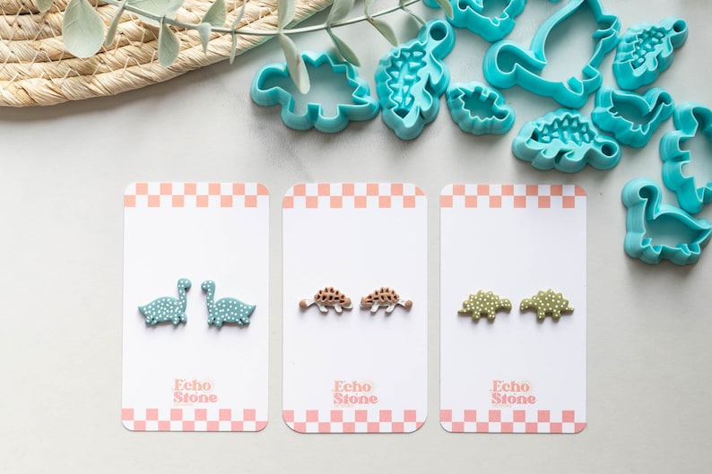 May include: Three cards with pairs of dinosaur-shaped polymer clay earrings. The earrings are on white cards with a pink and white checkered border. The text "Echo Stone" is printed on each card. The earrings are green, brown, and light green with white polka dots.