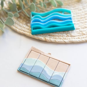 May include: A set of four silicone moulds for making soap or resin. The moulds are shaped like waves and have a light blue, teal, and white colour scheme. The moulds are sitting on a white surface.
