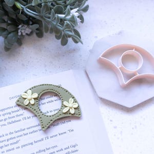 Puede incluir: Un marcapáginas verde y blanco con forma de abanico. El marcapáginas tiene un pequeño agujero en el centro y está decorado con flores blancas. El marcapáginas está sobre un libro con texto visible.