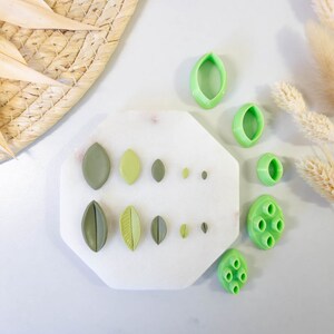 May include: A set of green plastic leaf-shaped cookie cutters on a white marble surface. The cutters are arranged in rows of different sizes.