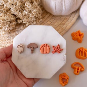 Puede incluir: Un conjunto de seis pendientes de arcilla con temática otoñal en varias formas, incluyendo una bellota, un hongo, una calabaza, una hoja y dos bellotas más. Los pendientes se muestran sobre una superficie de mármol blanco.
