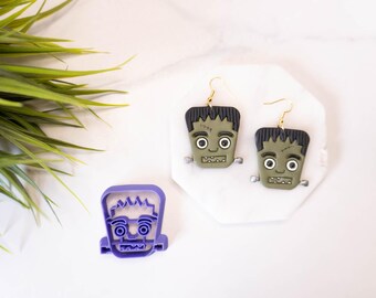 May include: A pair of green and black Frankenstein's monster earrings with gold hooks. A purple plastic cookie cutter of a Frankenstein's monster is also shown.