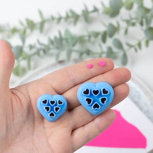 May include: Two blue heart-shaped cookie cutters with black heart cutouts. The larger cutter has seven cutouts and the smaller cutter has six cutouts.