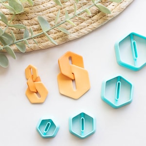 May include: Five cookie cutters, four are blue and one is orange. The blue cutters are hexagonal shaped with a small rectangle cut out in the center. The orange cutter is a more complex shape with two connected hexagons.