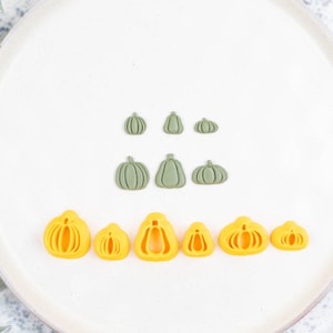 May include: A set of nine pumpkin-shaped cookie cutters, six in green and three in yellow. The cutters are arranged in two rows on a white plate.