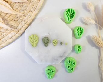May include: A set of six green leaf-shaped cookie cutters on a white marble surface. The cutters are different sizes and have a detailed leaf design.