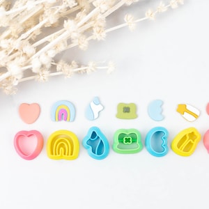 Może przedstawiać: Zestaw 12 kolorowych plastikowych foremek do ciastek w różnych kształtach, w tym serca, tęcze, chmury i koniczyny. Formy są ułożone w rzędzie na białym tle.