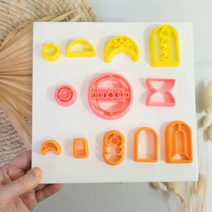May include: A set of 12 cookie cutters in various shapes and sizes. The cutters are made of plastic and are in shades of pink, orange, and yellow. The cutters are arranged on a white background.