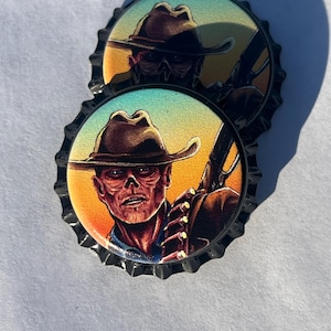 May include: Two metal bottle caps with a detailed illustration of a figure wearing a cowboy hat and holding a rifle. The artwork features a warm color palette with orange, yellow, and blue hues, set against a black, ridged edge.