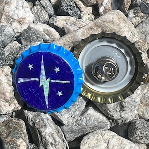 May include: A blue bottle cap pin with a white and green design featuring a lightning bolt and stars. The pin is set against a backdrop of gray rocks, with another bottle cap visible.