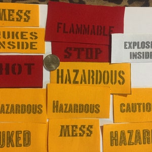 Puede incluir: Un conjunto de parches de tela en varios colores, incluyendo rojo, amarillo y blanco. Cada parche presenta texto con plantilla como "MESS", "HAZARDOUS" y "EXPLOSIVES INSIDE". Se incluye una moneda como referencia de tamaño.