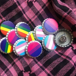 May include: A collection of colourful bottle caps featuring various pride flag designs, including the transgender, bisexual, and lesbian flags. The bottle caps are arranged in a circle on a pink and black checked fabric.
