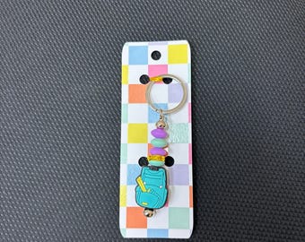 May include: A keychain featuring a teal backpack charm with a yellow ruler detail, attached to a silver ring. The charm is accented with purple, mint green, and yellow beads, and is displayed on a rectangular card with a multicolored checkerboard pattern.