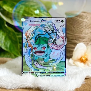 Puede incluir: Una carta de intercambio de Pokemon con un Bulbasaur VMAX verde con un símbolo Gigantamax. La carta tiene un fondo azul, rosa y blanco con un diseño de peces koi nadando alrededor de Bulbasaur. La carta tiene un borde verde y el texto "Bulbasaur VMAX" y "120 PS" en blanco.