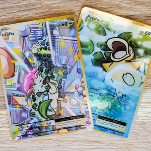 May include: Two Pokemon trading cards with gold borders. The card on the left is Leafia, a basic Pokemon with 60 HP. The card on the right is Psyduck, a basic Pokemon with 60 HP. Both cards feature colorful illustrations and text that includes the Pokemon's name, HP, and abilities. The Leafia card features a light pink and green Pokemon with a leafy tail. The Psyduck card features a yellow duck Pokemon with a blue background.