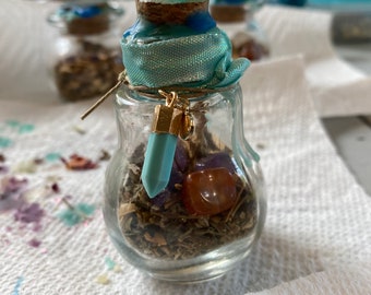 May include: A small glass jar with a cork lid and a blue ribbon tied around the top. The jar is filled with dried herbs and crystals, including a red carnelian, a purple amethyst, and a light blue crystal.