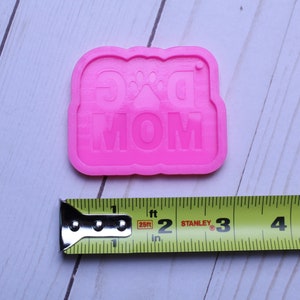 May include: Pink silicone mold for making a dog mom keychain. The mold is about 2 inches wide and 1.5 inches tall. The mold has the words "Dog Mom" and a paw print in the center.