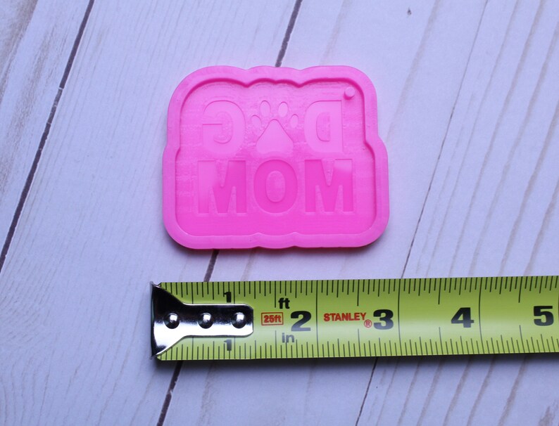 May include: Pink silicone mold for making a dog mom keychain. The mold is about 2 inches wide and 1.5 inches tall. The mold has the words "Dog Mom" and a paw print in the center.