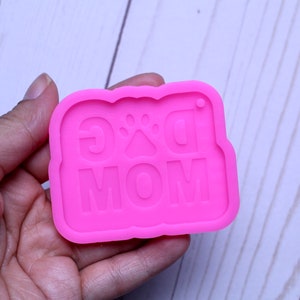 May include: Pink silicone mold for resin crafts. The mold is shaped like a banner with the words "Dog Mom" and a paw print in the center.