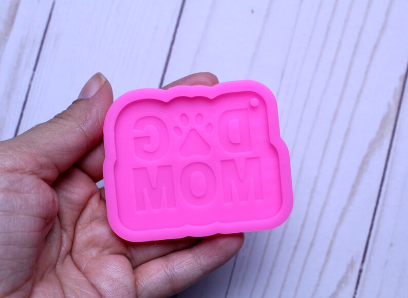 May include: Pink silicone mold for resin crafts. The mold is shaped like a banner with the words "Dog Mom" and a paw print in the center.