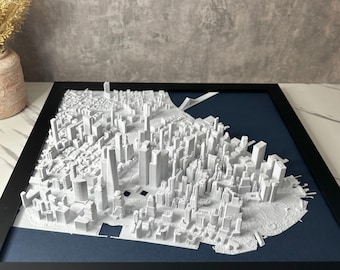 Puede incluir: Un modelo de ciudad en 3D impreso en blanco, enmarcado en un marco negro con un paspartú azul oscuro. El modelo es detallado y muestra los edificios y las calles de la ciudad.
