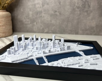 Puede incluir: Un modelo de ciudad en 3D impreso en blanco, en un marco negro con un paspartú azul oscuro. El modelo es detallado e incluye edificios, calles y un río.