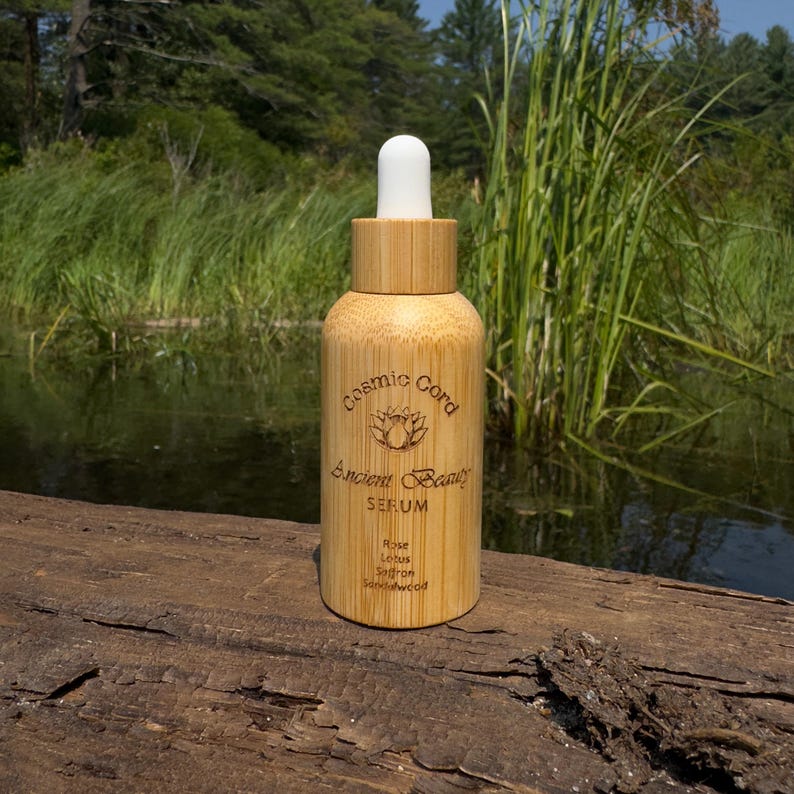 May include: A bamboo serum bottle with a white dropper. The bottle is cylindrical with the words "Cosmic Cord" and "Ancient Beauty" printed on it, along with the word "SERUM" and the ingredients: Rose, Lotus, Saffron, and Sandalwood.