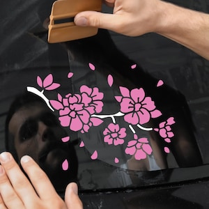 May include: A person is applying a pink floral decal to a black car window. The decal features a branch of cherry blossoms with white stems and pink flowers.