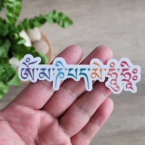 May include: A white sticker with the Tibetan phrase "Om Mani Padme Hum" written in colorful script. The letters are in a rainbow gradient, starting with purple and ending with red.