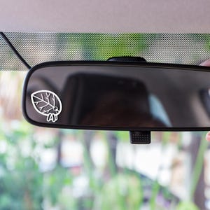 May include: A white leaf and insect sticker is attached to a car's rearview mirror. The sticker's design is a simple outline. The mirror reflects the car's interior and exterior surroundings.