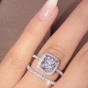 Puede incluir: Un anillo de plata con una piedra preciosa grande, de corte cuadrado, transparente, rodeada de piedras más pequeñas. El anillo se lleva en el dedo anular de una mano con uñas largas y manicuradas.