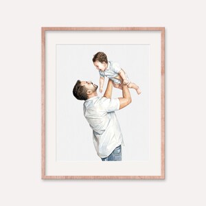 May include: A framed watercolor print depicts a father lifting a baby in the air. The father wears a white shirt and blue jeans. The baby is in a white onesie. The frame is a light brown color.
