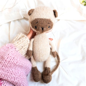 May include: A hand-knitted plush toy cat with brown and beige fur. The cat has a long tail and is sitting on a white surface.