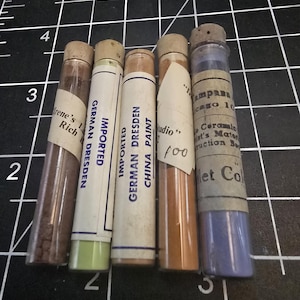 Könnte beinhalten: Fünf kleine Glasröhrchen mit Korkstopfen, die jeweils ein anderes farbiges Pulver enthalten. Die Etiketten auf den Röhrchen lauten "Importierte Deutsche Dresdner Farbe", "China Farbe", "Studio", "Irene's Rich" und "Violette Farbe".