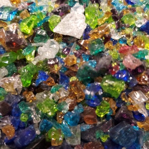 Könnte beinhalten: Nahaufnahme einer Sammlung farbenfroher, unregelmäßig geformter Glasstücke. Die Glasscherben sind in verschiedenen Blau-, Grün-, Bernstein-, Weiß- und Lilatönen gehalten. Die Stücke sind verstreut und erzeugen eine strukturierte und lebendige Darstellung.