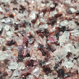 May include: A close-up of a mixture of clear and iridescent pink and purple glass pieces. The glass pieces are small and irregular in shape, and they are scattered randomly across the surface.