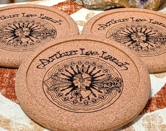 Puede incluir: Tres posavasos redondos de corcho con el texto "Arthur Lee Land" y un diseño gráfico central. Los posavasos son de color marrón claro y descansan sobre una superficie estampada, con una amatista púrpura y un cristal en el fondo.