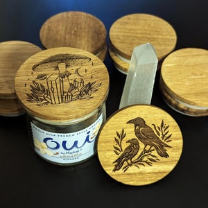 May include: A set of six wooden candle lids with various designs. The lids feature a mushroom with crystals, two ravens, and abstract patterns. The lids are made of natural wood and have a rustic look. The lids are perfect for adding a touch of whimsy to your home decor.
