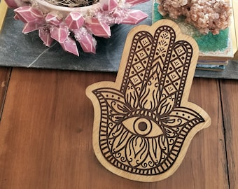 Può includere: Un ornamento in legno a forma di mano con intagli intricati e un grande occhio al centro. L'ornamento è marrone e ha una venatura del legno chiara. È un simbolo tradizionale di protezione e buona fortuna.