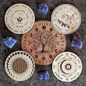 Puede incluir: Una colección de tableros de adivinación de madera con varios diseños, incluyendo un árbol de la vida, un lobo y patrones geométricos. Cada tablero presenta texto como "Sí", "Tal vez" y "Reformular". Cristales de amatista están dispersos alrededor.