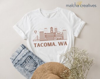 May include: White t-shirt with a brown Tacoma, WA skyline print. The design shows buildings, a bridge, and a hot air balloon. The shirt is styled with denim shorts and a woven straw hat.