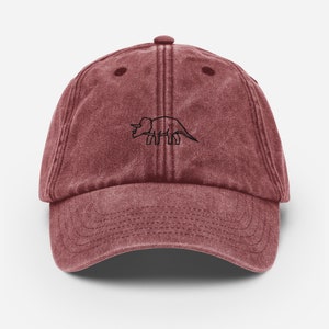 May include: A maroon vintage-style baseball cap with a black outline of a triceratops dinosaur on the front.
