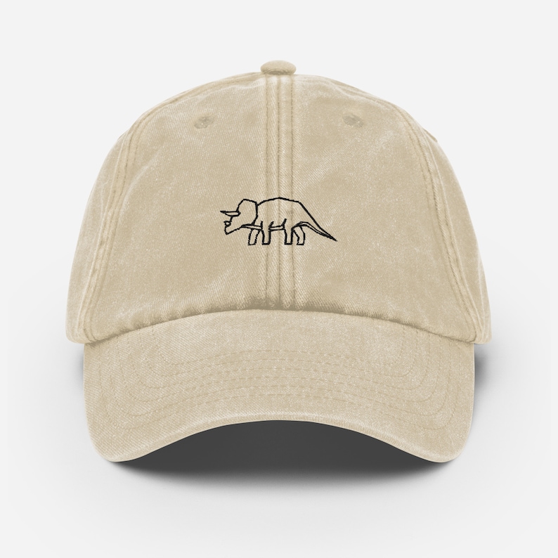 May include: A beige cotton baseball cap with a black embroidered outline of a triceratops dinosaur.