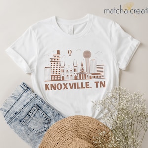 Puede incluir: Camiseta blanca con un contorno marrón del horizonte de Knoxville, TN y el texto "KNOXVILLE, TN".