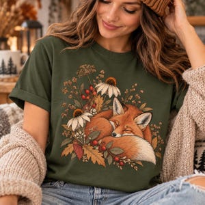 Peut inclure: Un t-shirt vert olive arborant une illustration graphique d'un renard orange endormi, recroquevillé au milieu d'un feuillage d'automne composé de fleurs blanches, de baies rouges et de feuilles de chêne brunes, centré sur le devant.