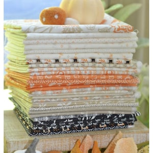 May include: A stack of folded fabric in various patterns and colors, including orange, cream, and black. A small pumpkin and decorative felt balls sit atop the stack. The fabrics appear to be suitable for quilting or other craft projects.