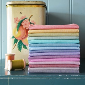 May include: A stack of 14 fabric squares in various pastel colors, including pink, purple, blue, green, and yellow. The fabric squares are stacked on a dark blue surface, with two spools of thread in front of them.